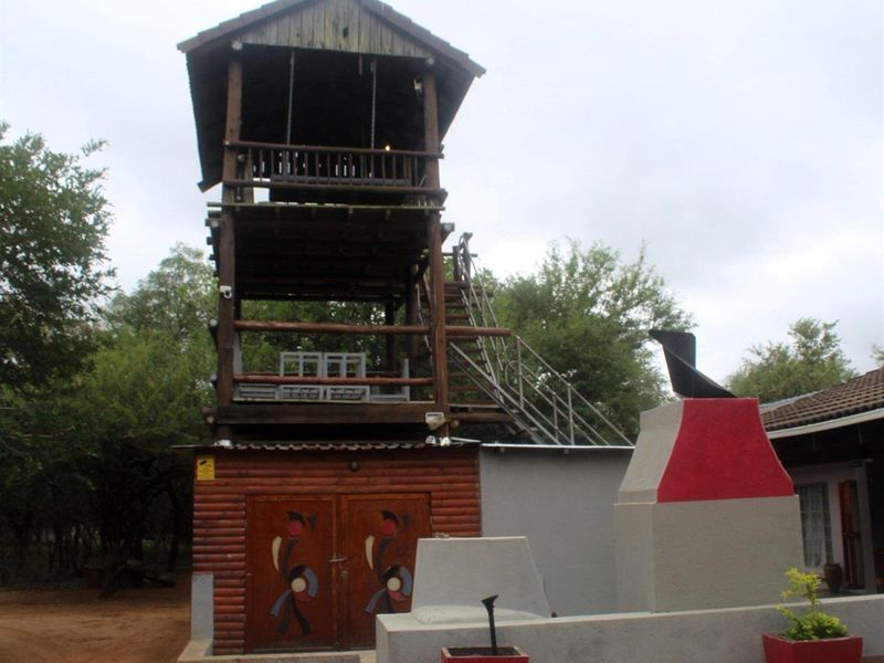 A multi-level wooden observation tower structure featuring an external staircase and a built-in outdoor braai area in a natural garden setting.