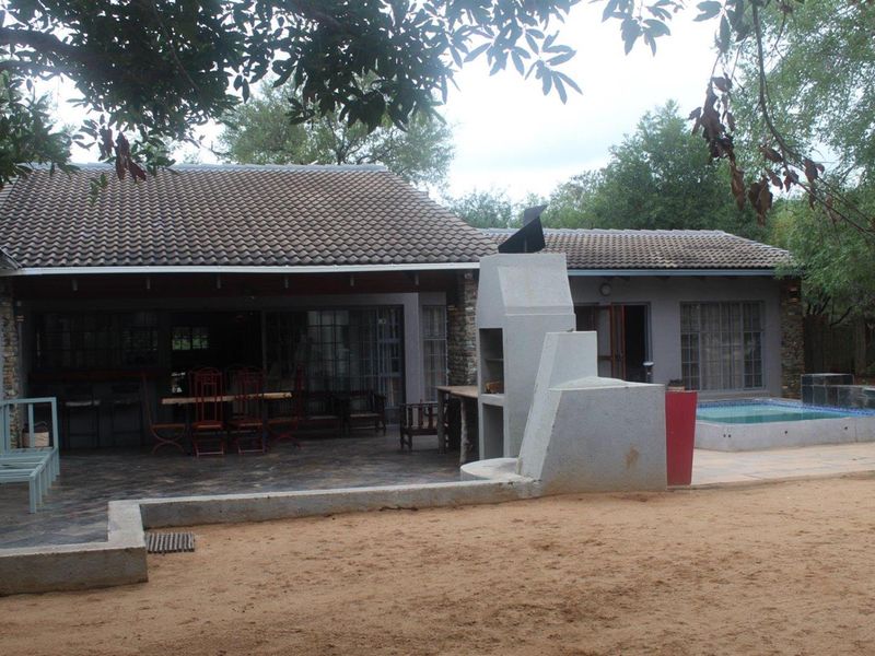 Rear view of the property featuring a covered patio, built-in braai area, and a small swimming pool set within a natural garden environment.