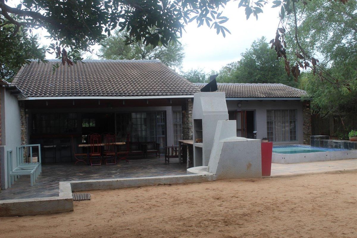 Rear view of the property featuring a covered patio, built-in braai area, and a small swimming pool set within a natural garden environment.