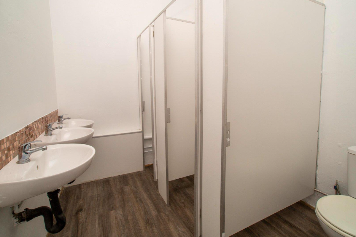 Commercial restroom facility featuring multiple wall-mounted washbasins with mosaic tile splashbacks and partitioned toilet cubicles with wood-look vinyl flooring.
