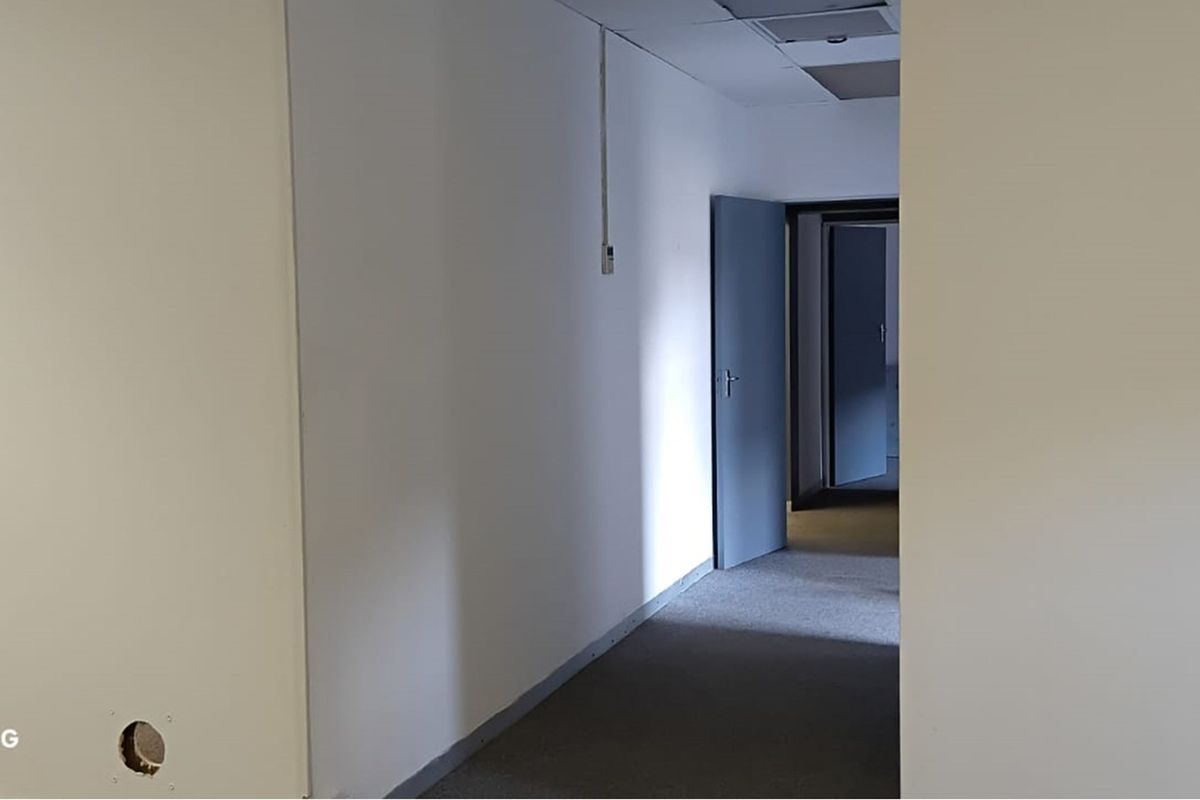 A vacant office hallway featuring carpet flooring, suspended ceiling tiles, and multiple internal doors, currently showing signs of minor wall damage and exposed wiring.