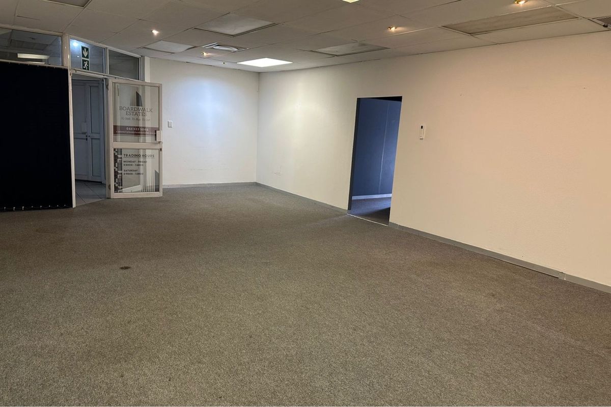 An empty, open-plan office space featuring a suspended ceiling, recessed lighting, and carpeted flooring, with a glass entrance door and internal doorway.