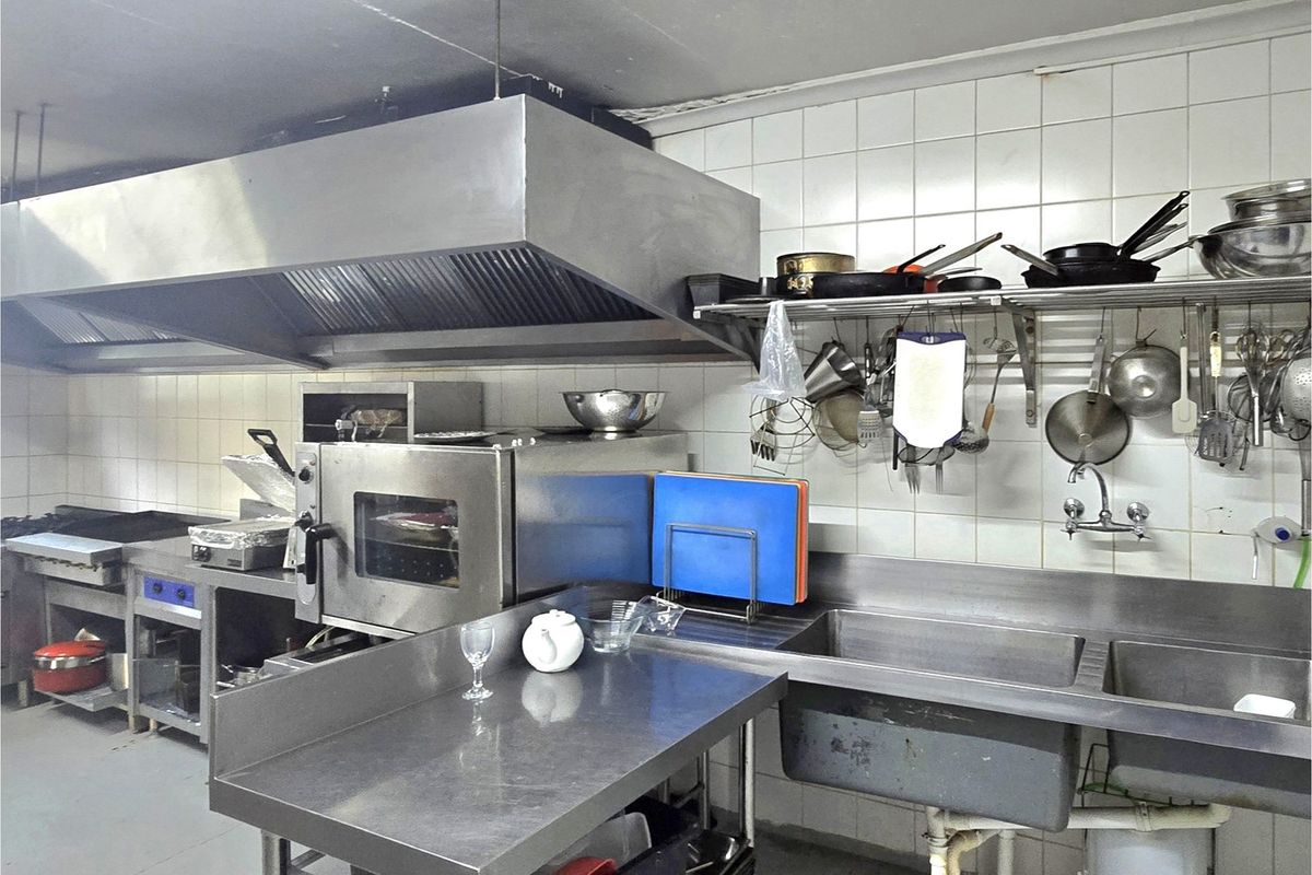 Commercial-style kitchen featuring stainless steel work surfaces, a large industrial extractor hood, double sink, and tiled wall splashbacks.