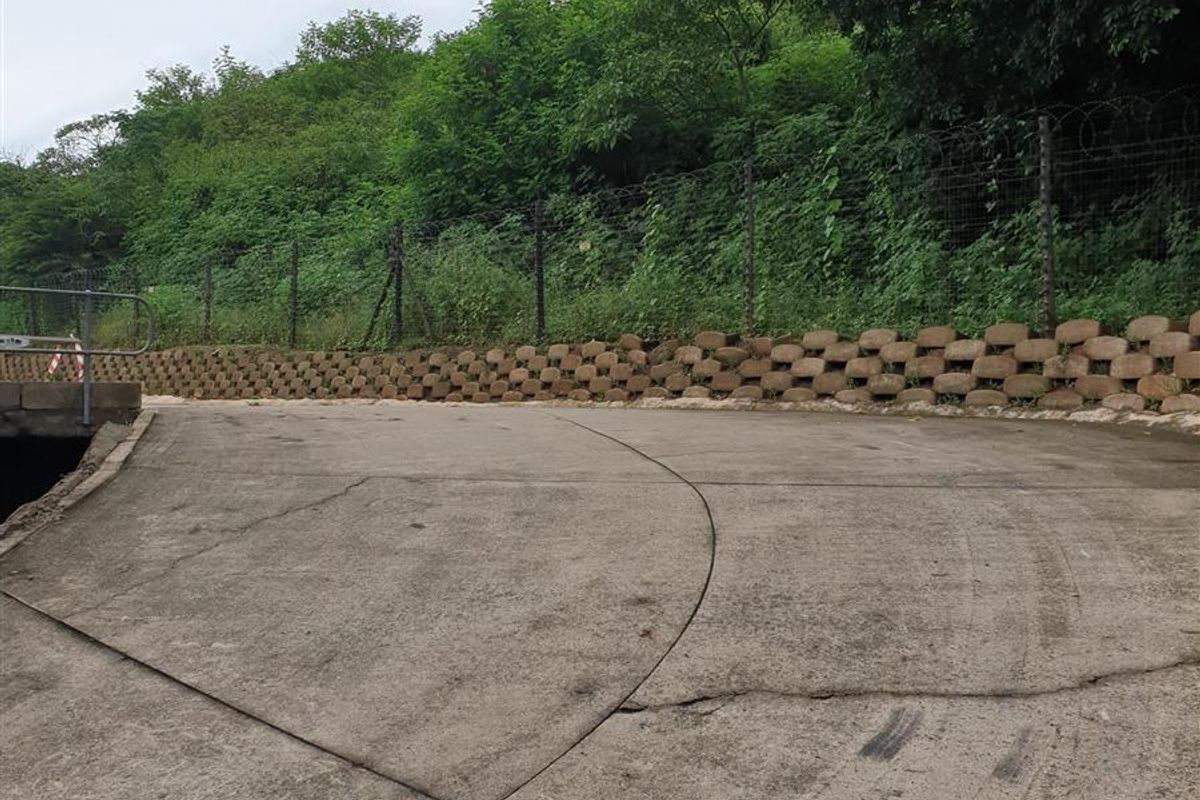 An industrial outdoor area featuring a concrete driveway, retaining wall, and perimeter security fencing with razor wire.