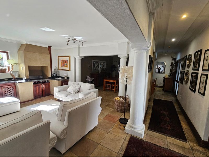 An open-plan living area featuring a built-in indoor braai, classic architectural columns, and natural stone flooring, seamlessly connecting to the dining space and hallway.