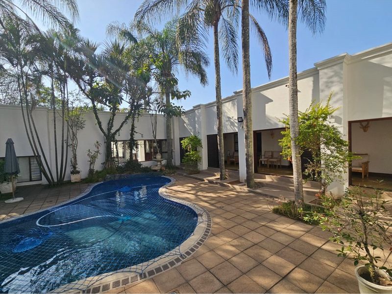 A tranquil courtyard featuring a kidney-shaped swimming pool with a safety net, surrounded by paved patio areas and mature palm trees.