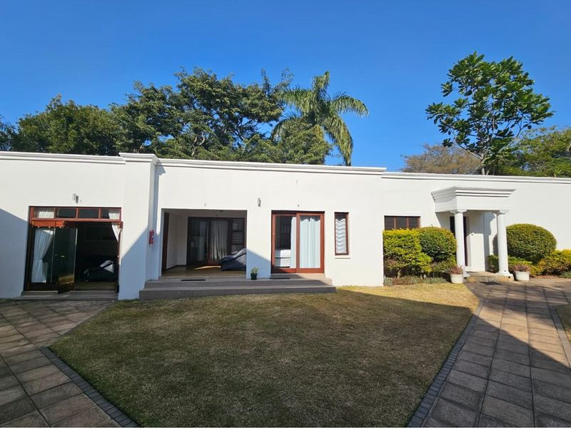 A well-maintained rear exterior view of the guest house featuring a paved courtyard, manicured lawn, and multiple access points with sliding glass doors.