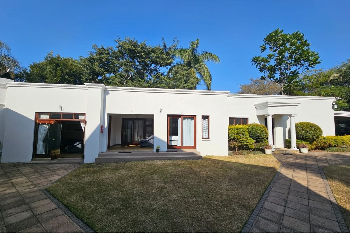 A well-maintained rear exterior view of the guest house featuring a paved courtyard, manicured lawn, and multiple access points with sliding glass doors.
