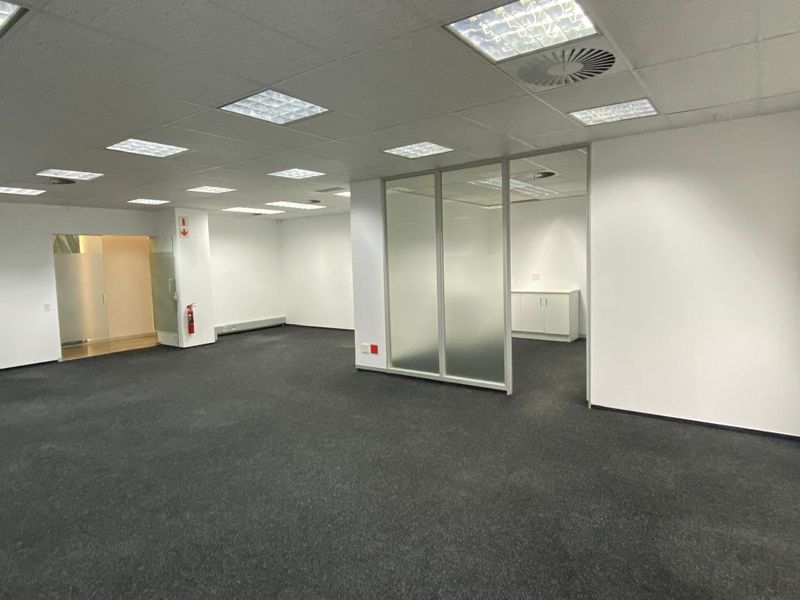 Open-plan office space featuring a suspended ceiling with recessed lighting, carpeted flooring, and a glass-partitioned private office area.