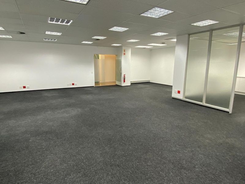 Spacious open-plan office area featuring a suspended ceiling with recessed lighting, glass partitioning, and wall-mounted electrical outlets.