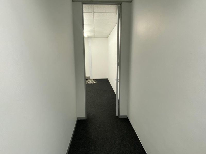 A narrow office hallway leading into an empty workspace featuring dark carpeting and a suspended acoustic tile ceiling.