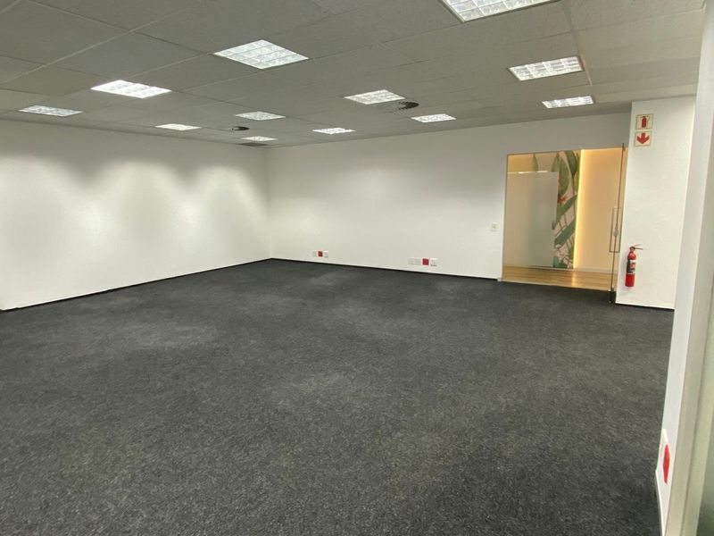 A spacious, open-plan office area featuring a suspended grid ceiling with recessed lighting, dark carpeting, and a glass-paneled doorway.