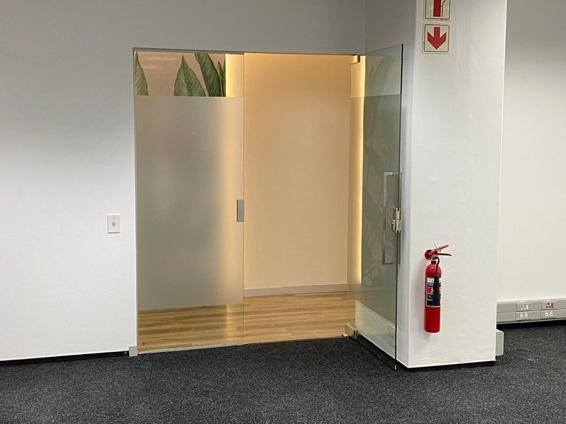 A modern office entrance featuring a frameless glass door with frosted detailing, leading into a bright, wood-floored workspace.
