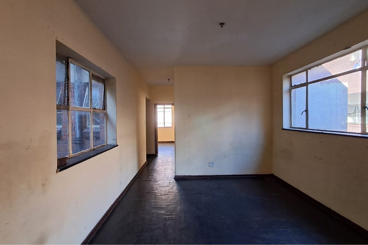 An empty, dated interior space featuring dark parquet flooring and large windows, requiring cosmetic updates and refurbishment.