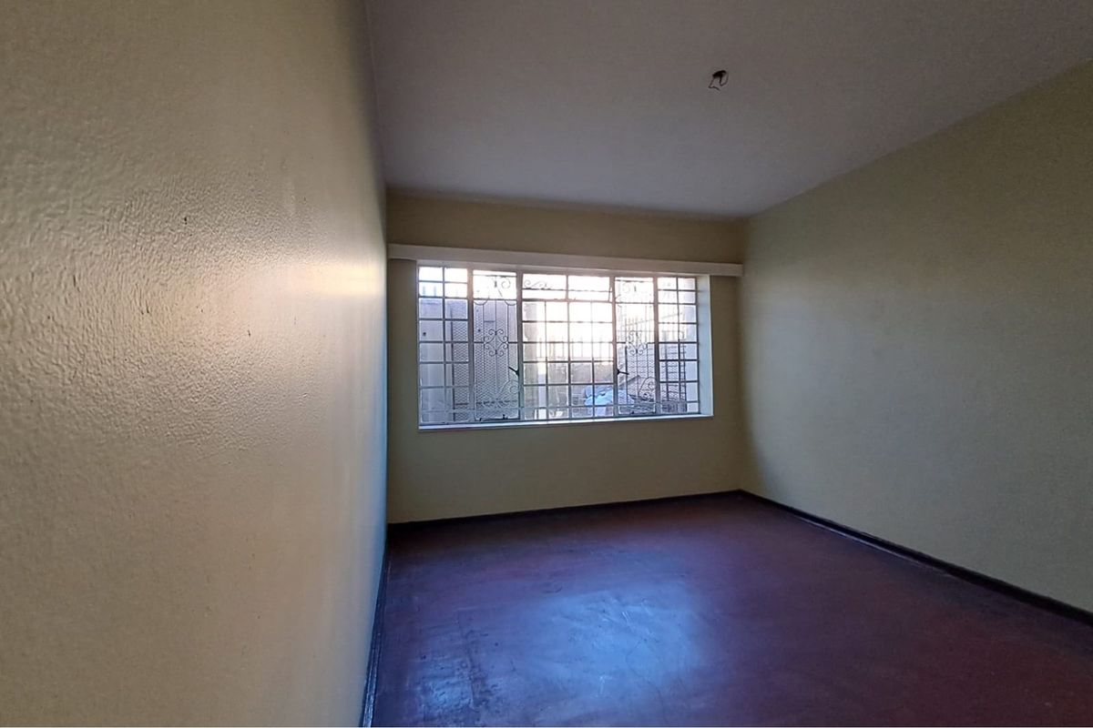 An empty bedroom featuring a large window with security bars and worn flooring, awaiting renovation or staging to showcase its potential.