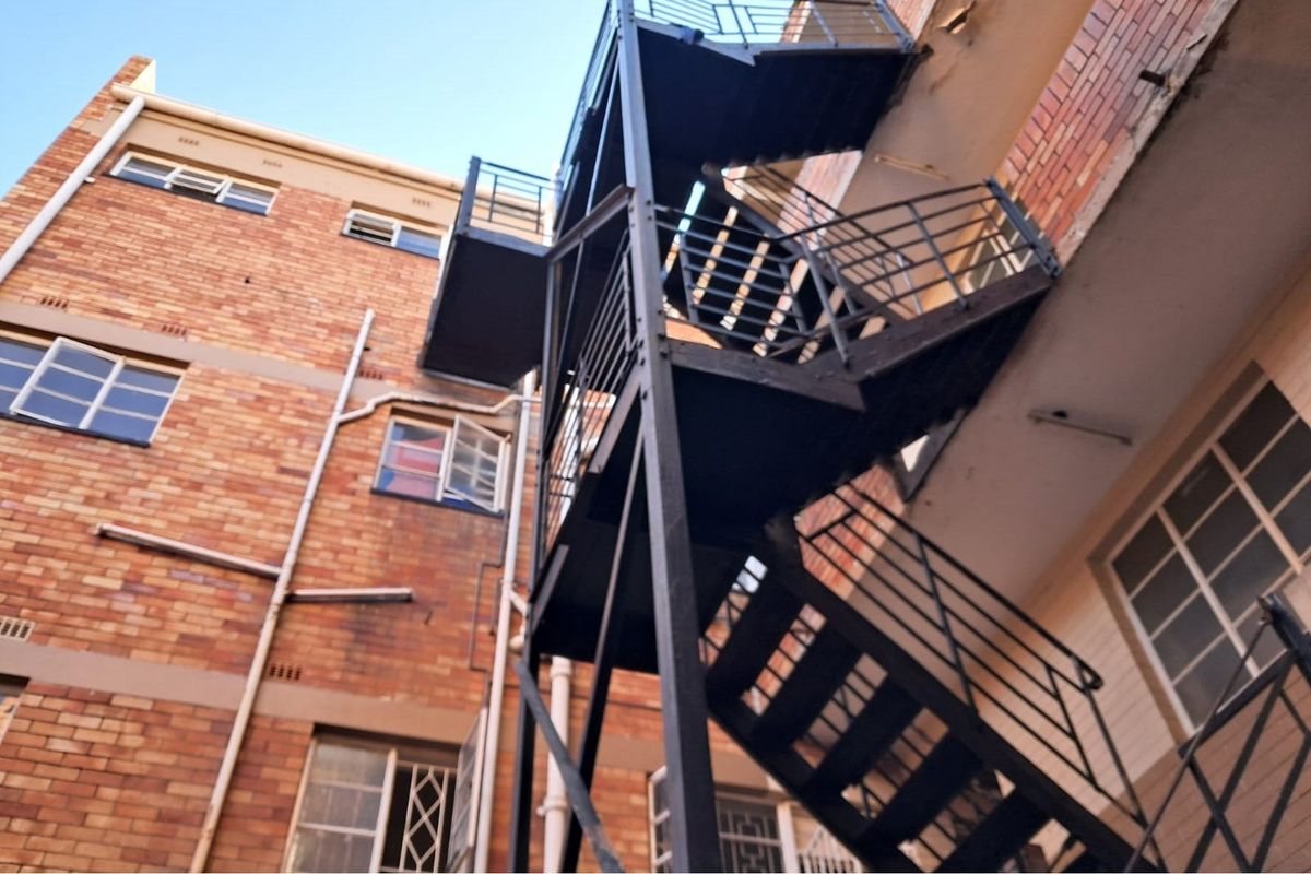 Exterior view of a multi-story brick building featuring an external metal fire escape staircase and steel-framed windows.