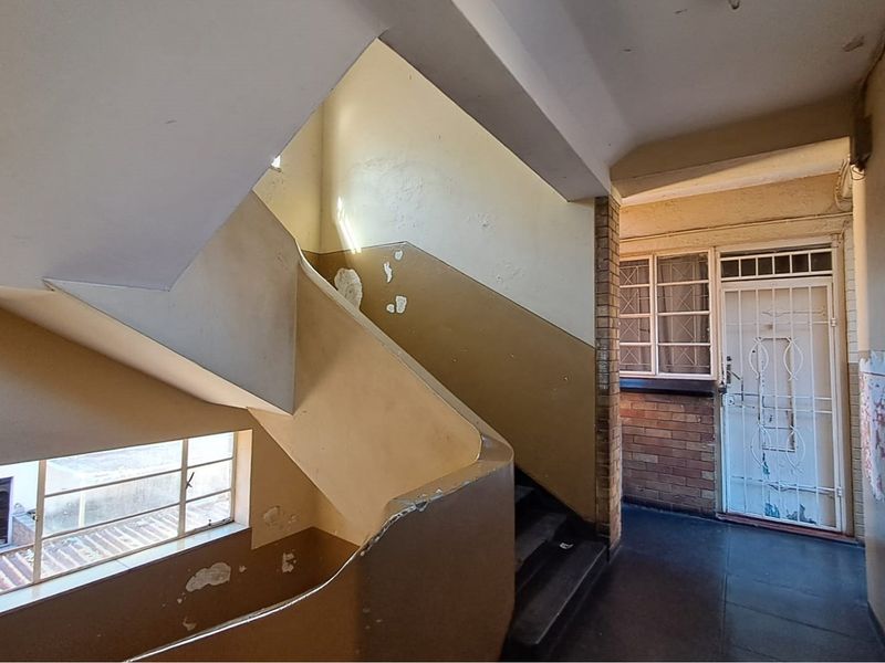 A dated interior hallway and staircase area featuring brick accents, steel-framed windows, and a security gate, currently showing significant wear to wall finishes.