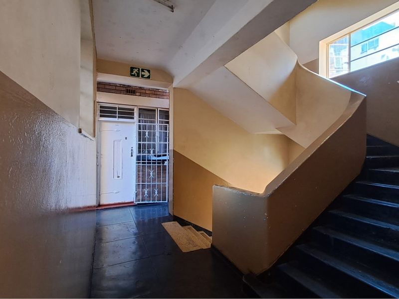 A functional interior hallway and stairwell area featuring tiled flooring, a security gate, and emergency signage, showing signs of wear and dated finishes.