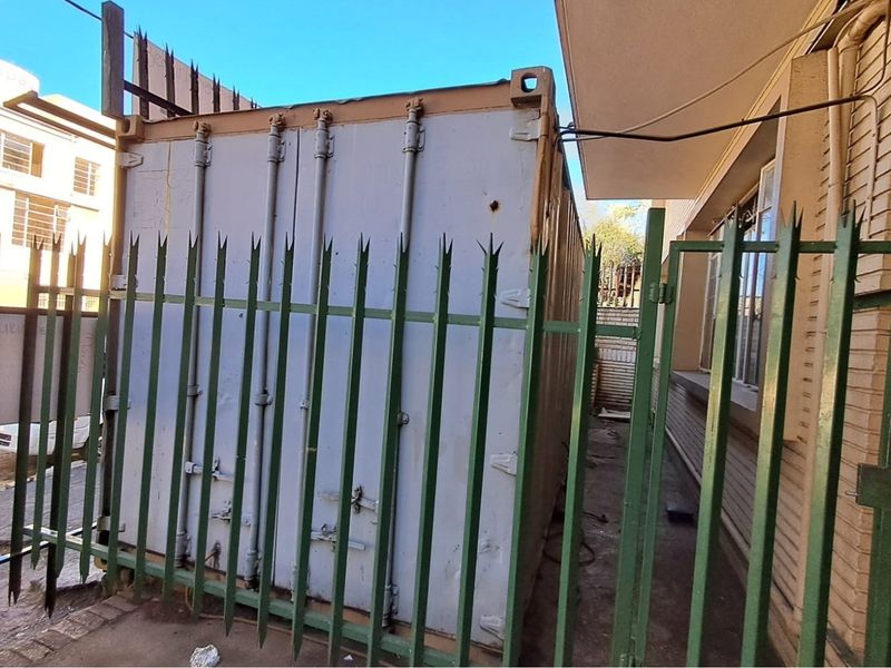 Exterior view of a side passage featuring a shipping container secured behind metal fencing and spikes adjacent to a brick building.