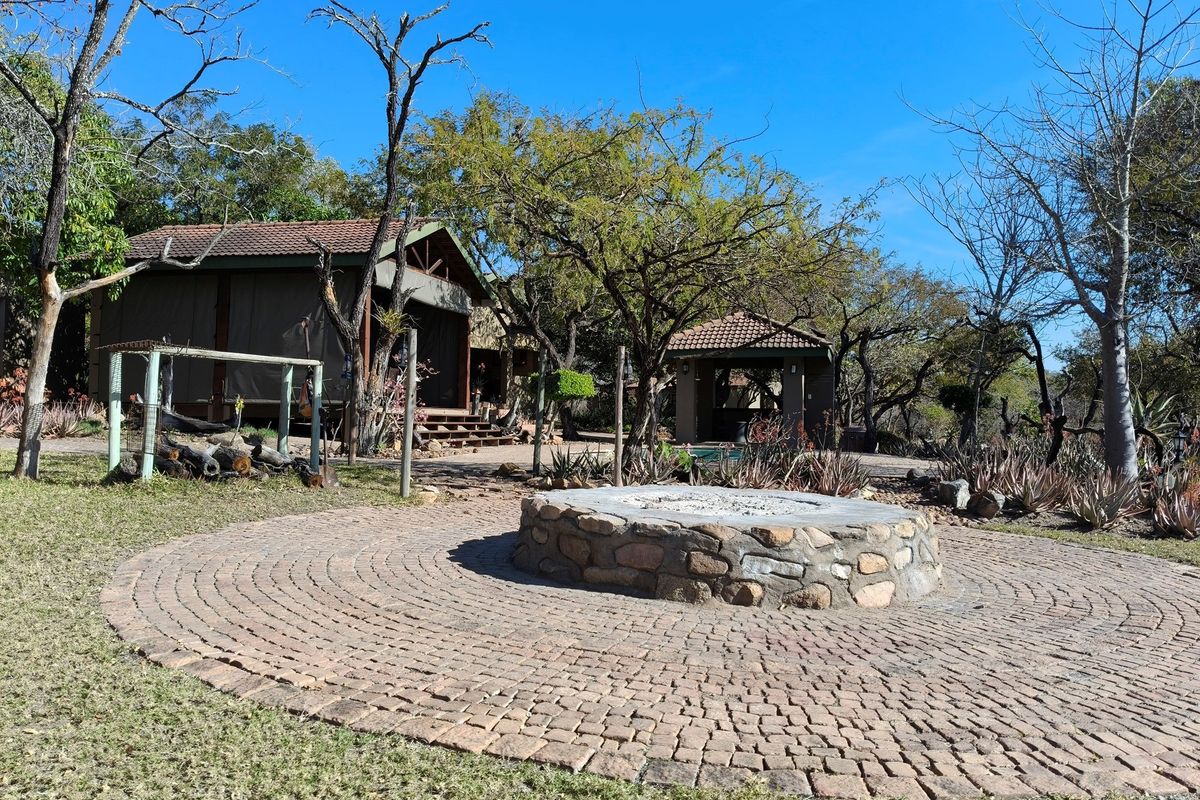 Outdoor boma area featuring a circular stone fire pit set within a paved courtyard, surrounded by landscaped gardens and lodge structures.