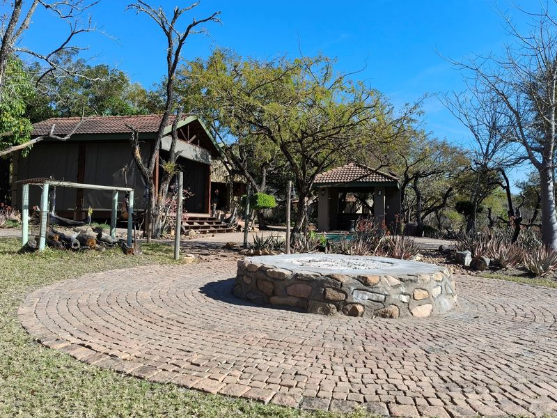 Outdoor boma area featuring a circular stone fire pit set within a paved courtyard, surrounded by landscaped gardens and lodge structures.