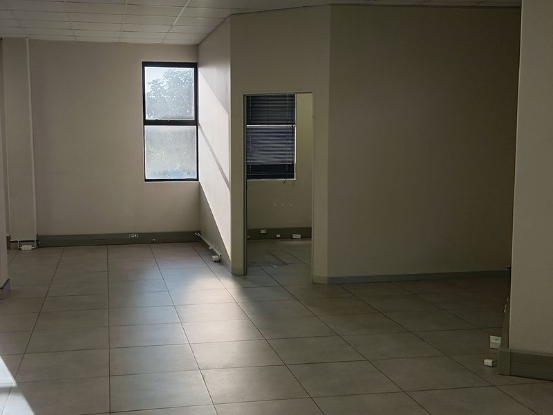 An empty, open-plan office space featuring neutral tiled flooring, a suspended ceiling, and natural light through aluminium-framed windows.