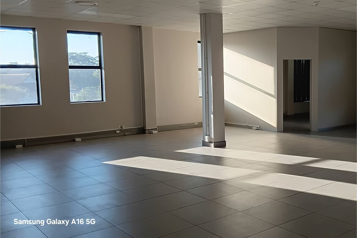 A spacious, empty open-plan office area featuring tiled flooring, a suspended ceiling, and natural light from large windows.