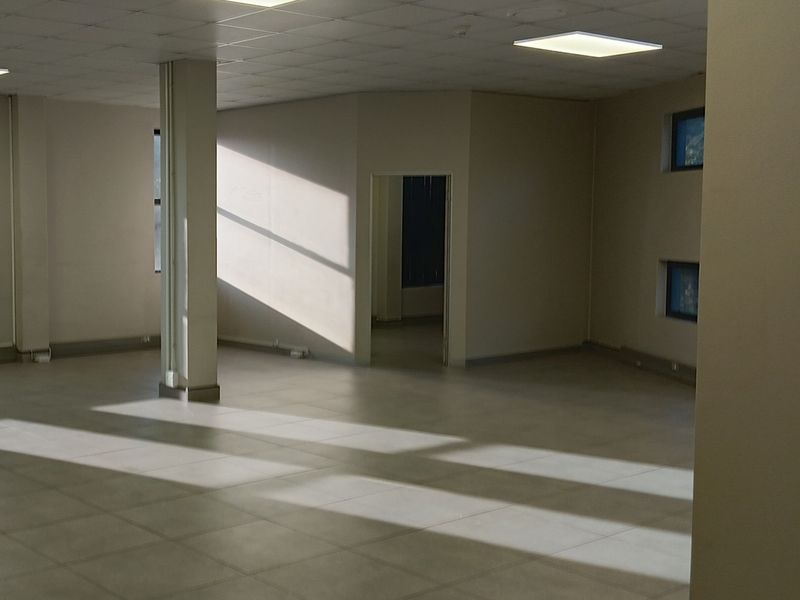 A spacious, vacant open-plan office area featuring neutral tiled flooring, a suspended ceiling with recessed lighting, and a central structural support column.