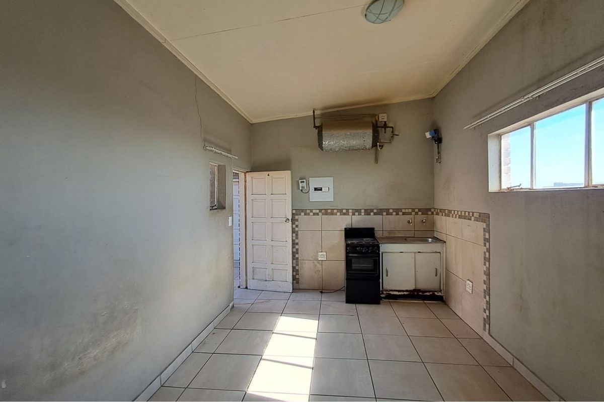 A basic kitchen area featuring tiled flooring, a small sink unit, a freestanding stove, and exposed wall-mounted water heating infrastructure.