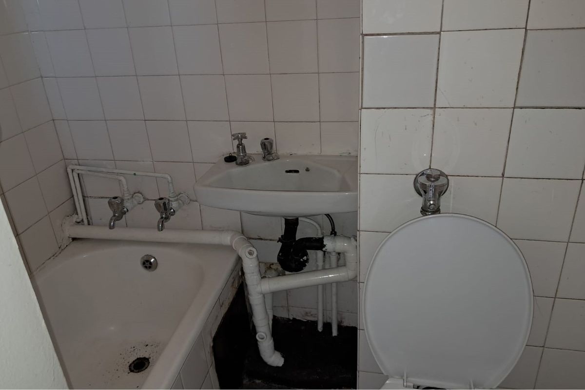 A compact bathroom featuring a small bathtub, wall-mounted basin, and toilet with visible exposed plumbing and dated wall tiling.