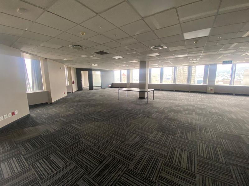 A spacious, open-plan office area featuring large windows with city views, suspended ceilings, and durable carpet tiles, offering a versatile workspace.