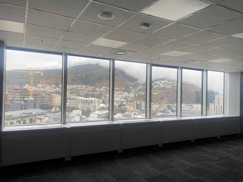 A bright, empty office space featuring large floor-to-ceiling windows with expansive city and mountain views, suspended ceilings, and dark carpet tiles.