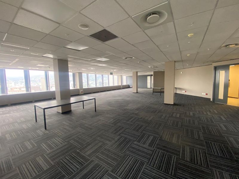 A spacious, open-plan office area featuring carpet tiles, suspended ceilings with integrated lighting, and large windows offering expansive city views.