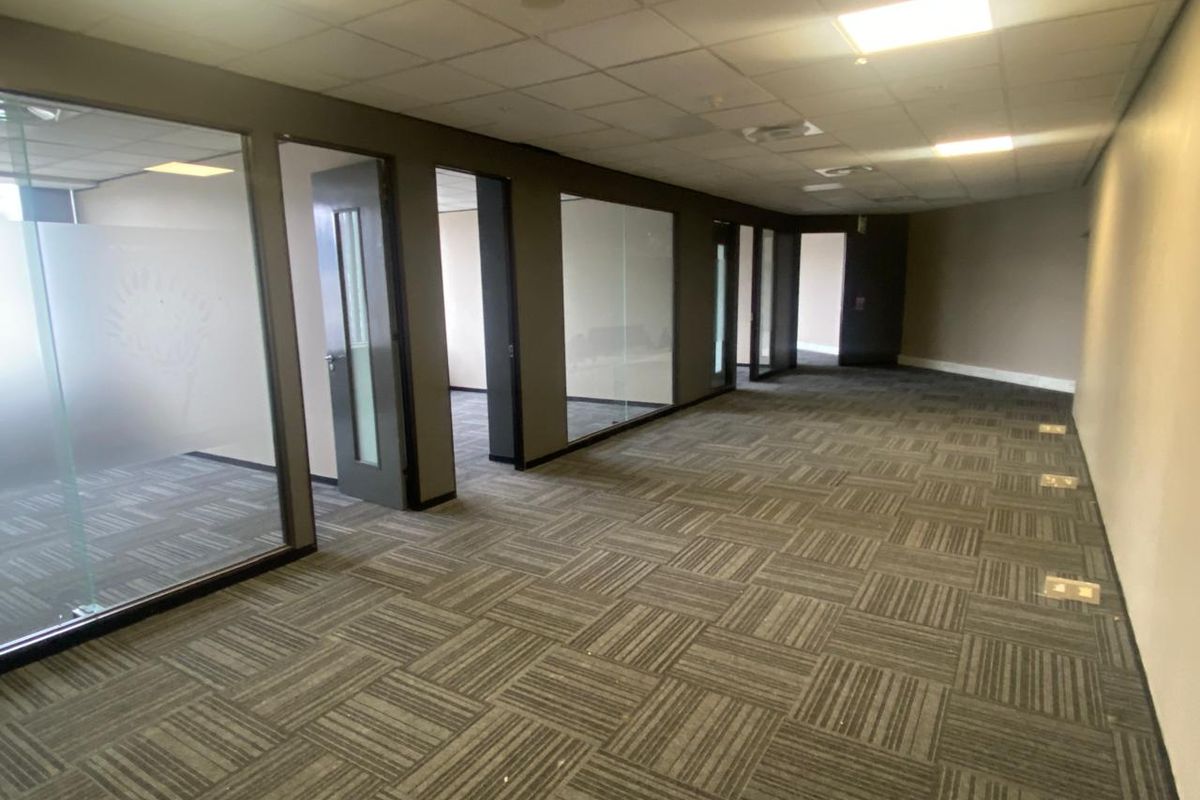 A spacious, vacant office area featuring modern glass partitions, carpet tile flooring, and a suspended ceiling with recessed lighting.