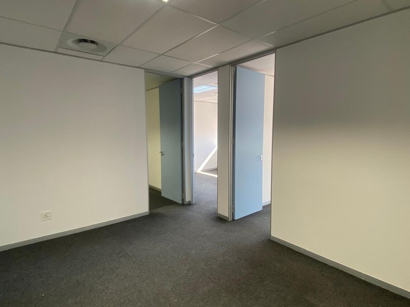 An empty office space featuring a suspended ceiling, recessed air conditioning, and carpeted flooring with access to adjacent rooms.