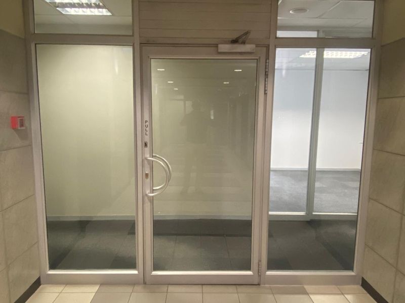 A clean, professional office entrance featuring a glass door with aluminum framing, tiled flooring, and recessed ceiling lighting.
