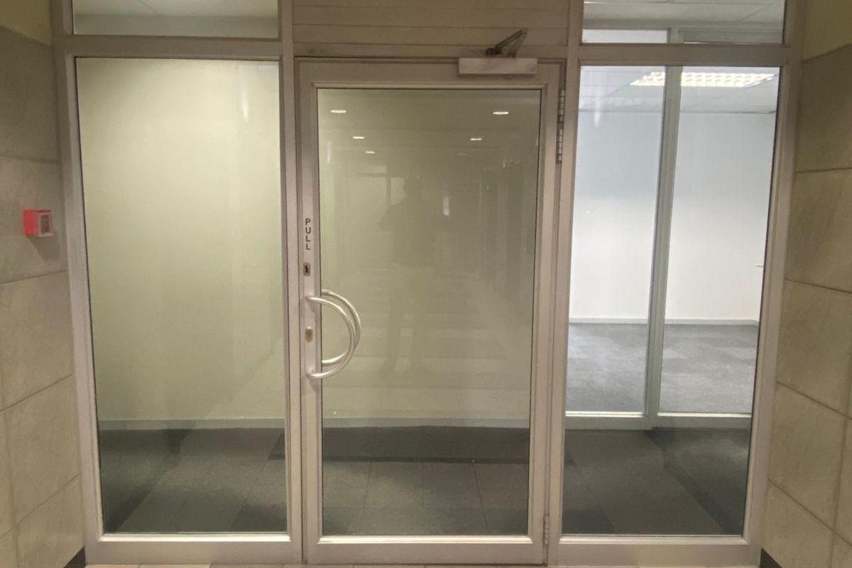 A clean, professional office entrance featuring a glass door with aluminum framing, tiled flooring, and recessed ceiling lighting.