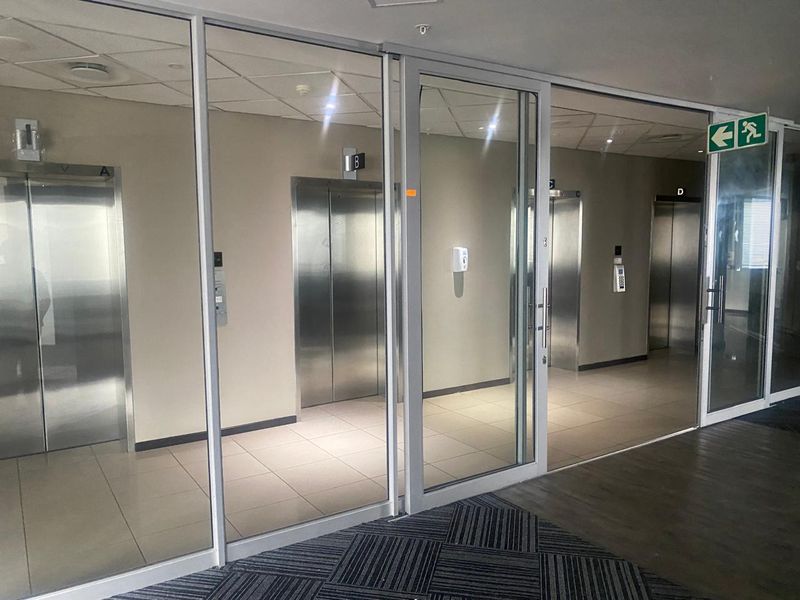 A modern office building elevator lobby featuring glass partitions, stainless steel elevator doors, and clean tiled flooring with professional lighting.