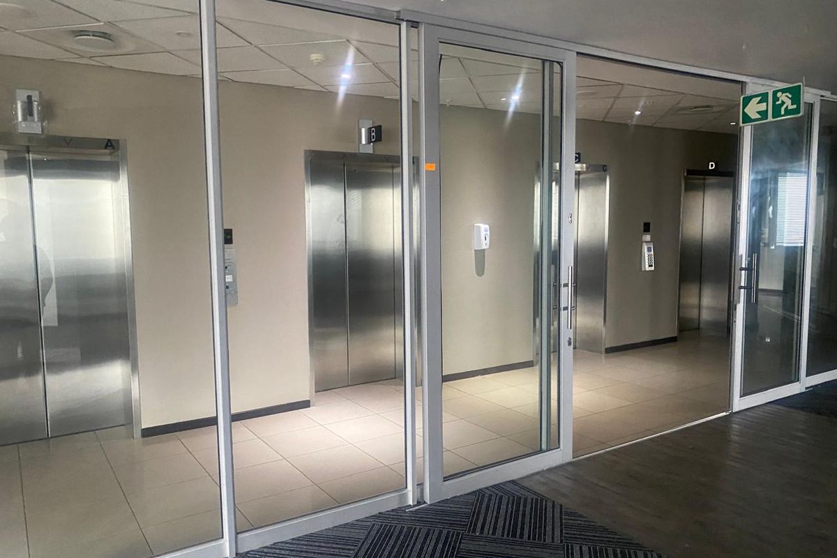 A modern office building elevator lobby featuring glass partitions, stainless steel elevator doors, and clean tiled flooring with professional lighting.