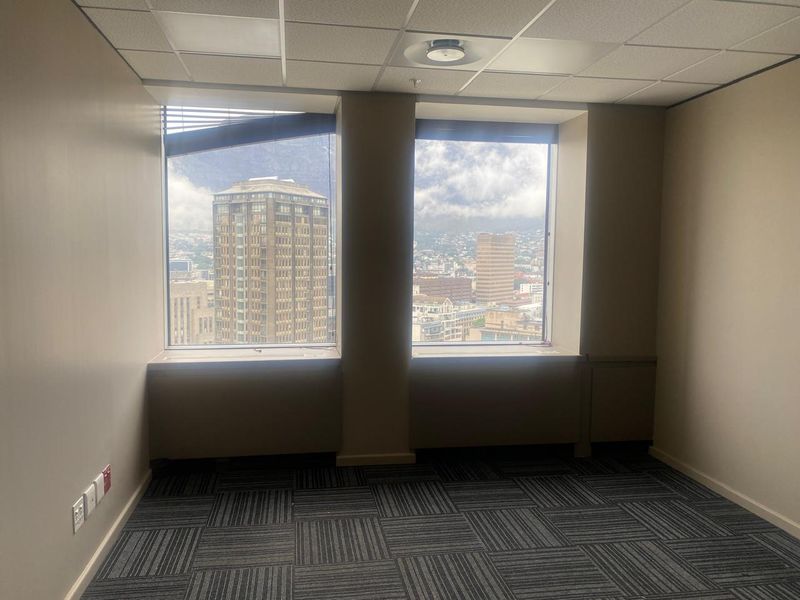 An empty office space featuring carpet tiles, a suspended ceiling, and large windows offering expansive city views.