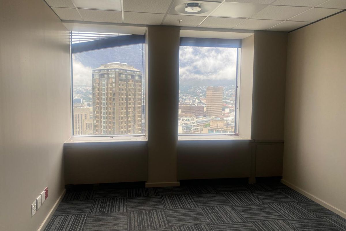An empty office space featuring carpet tiles, a suspended ceiling, and large windows offering expansive city views.