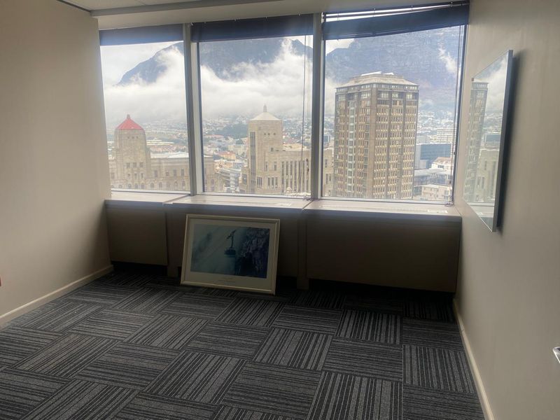 A bright, empty office space featuring large windows with expansive city and mountain views, neutral walls, and modern carpet tiles.