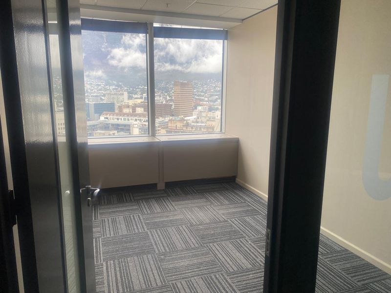 A bright, empty office space featuring carpet tiles, a suspended ceiling, and large windows offering expansive city views.