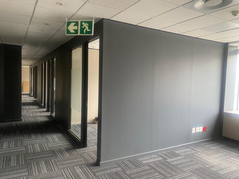 A modern office hallway featuring carpet tiles, suspended ceiling, and glass-partitioned office entrances with clear emergency signage.