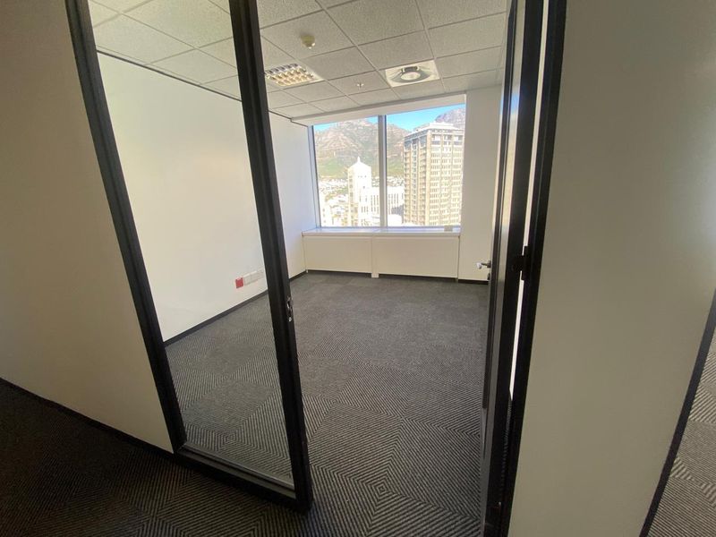 A bright, empty office space featuring carpeted flooring, a suspended ceiling, and a large window offering expansive city and mountain views.