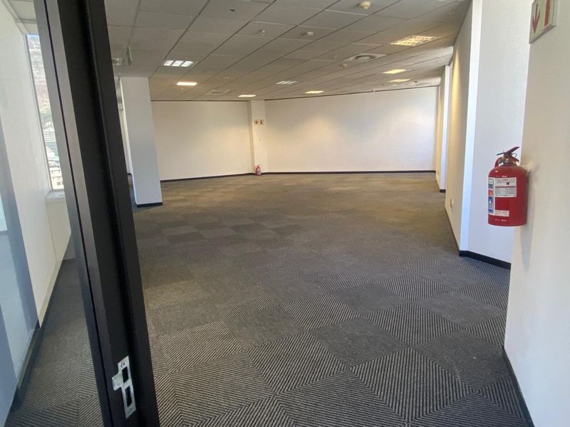 A spacious, open-plan office area featuring carpeted flooring, a suspended ceiling with recessed lighting, and integrated fire safety equipment.