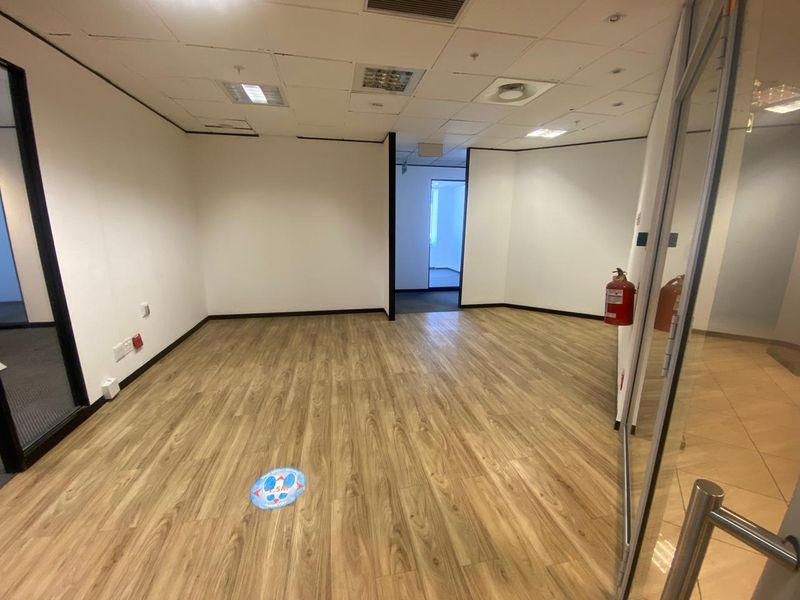 An empty, open-plan office space featuring wood-look laminate flooring, a suspended ceiling with recessed lighting, and a glass partition entrance.