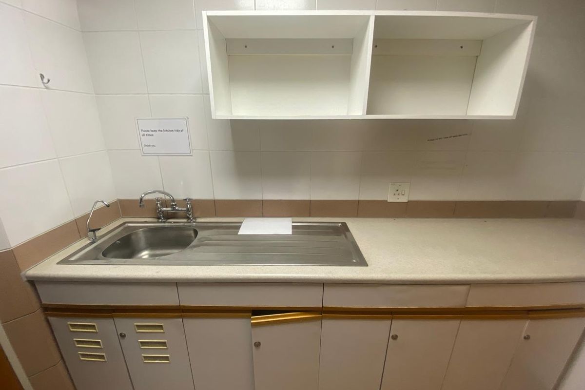 A small office kitchenette featuring a stainless steel sink, laminate countertop, and white cabinetry with wall-mounted storage shelves.