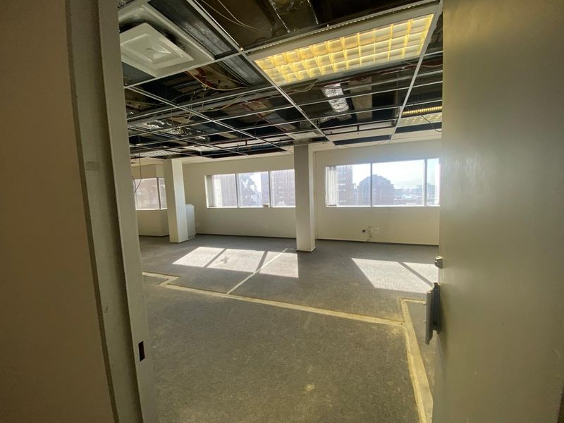 An empty office space featuring large windows, structural columns, and an exposed ceiling grid, currently requiring renovation and ceiling tile installation.