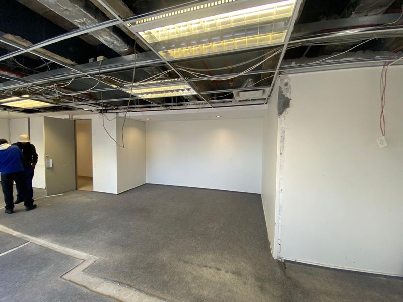 An empty office space undergoing renovation, featuring a partially exposed suspended ceiling, carpeted flooring, and unfinished wall surfaces.
